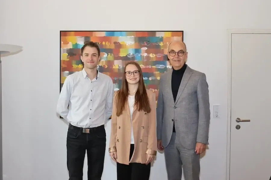 Drei Personen stehen für ein offizielles Foto nebeneinander, darunter eine junge Absolventin des nichttechnischen Verwaltungsdienstes, flankiert von zwei Vertretern der Regierung von Unterfranken vor einem farbigen Kunstwerk.