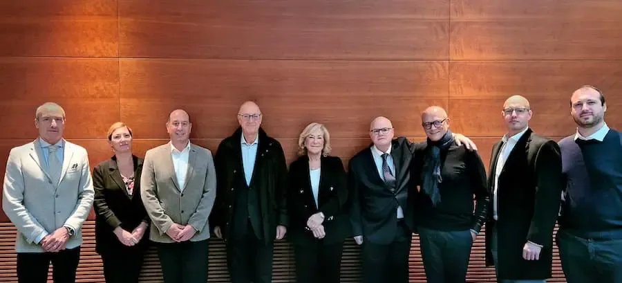 Gruppenfoto mehrerer Geschäftsleute während der Unterzeichnung einer strategischen Partnerschaft zwischen UNICOR und Olmas vor Holzwand.