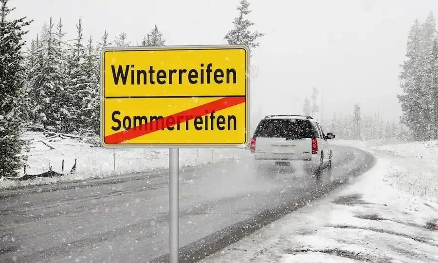 Ein Auto fährt bei Schneefall auf winterlicher Straße an einem Verkehrsschild vorbei, das den Wechsel von Sommer- auf Winterreifen anzeigt