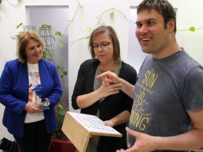 (Foto: Reto Glemser): Hilfsmittel aus der gestützten Kommunikation stärken die Fähigkeit, sich auszudrücken: Tobias Schimmel begrüßt damit die CSU-Landtagsabgeordnete Martina Gießübel (links), die von Gruppenleiterin Sandra Thein durch die Schweinfurter Tagesförderstätte für Menschen mit Autismus geführt wird. (Foto: Reto Glemser)